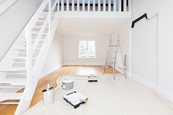 Bright interior space under renovation with white walls, wood floor, and ladder. Paint supplies and staircase visible.