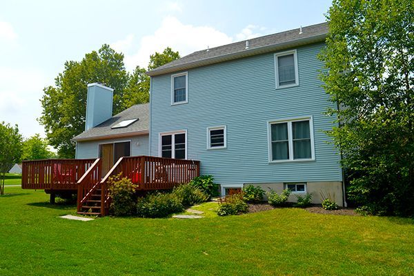 Backyard of a two-story blue house with a wooden deck, surrounded by green grass and trees.