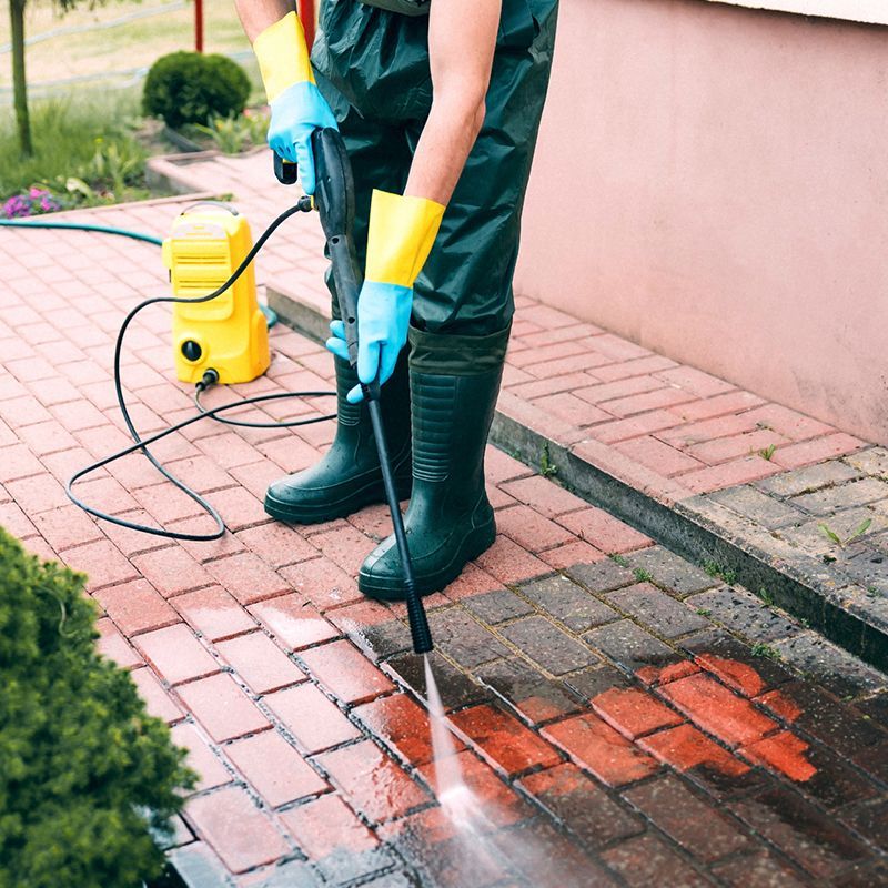 Person power washing red brick pavers. They wear green boots and rain pants, blue gloves, and yellow gloves.