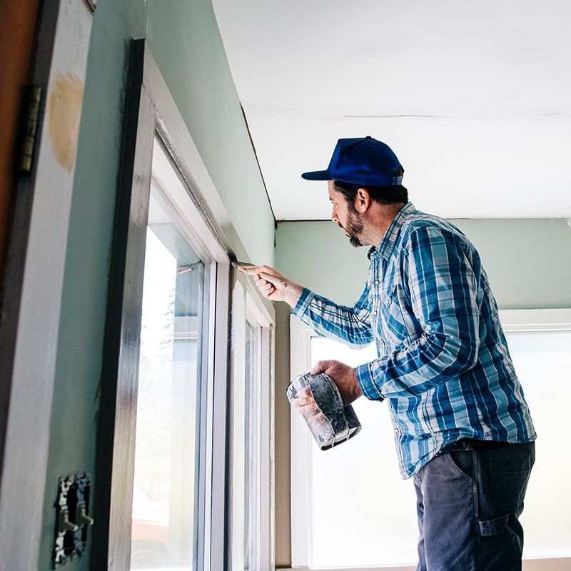 Man in blue plaid shirt paints window trim. Man in blue plaid shirt paints window trim.