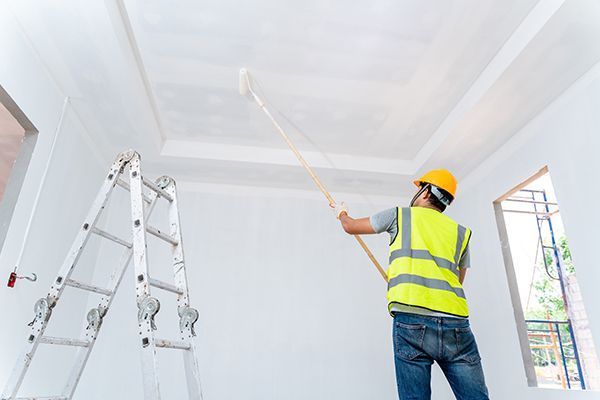 Painter in yellow safety vest and hard hat using roller on ceiling.