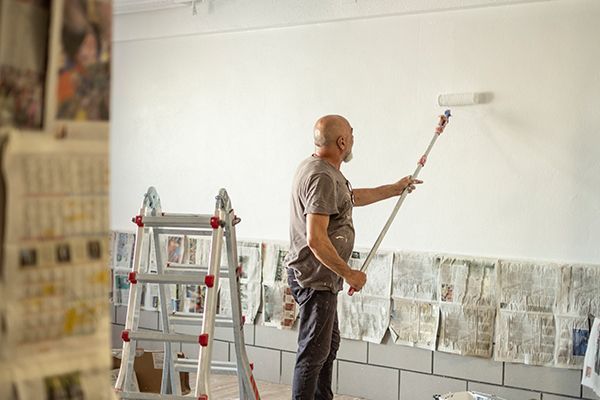 Man painting a white wall with a roller; newspapers protect the base, a ladder stands nearby.