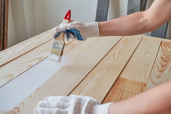 Person painting white paint on a light-colored wooden surface with a brush, wearing gloves.
