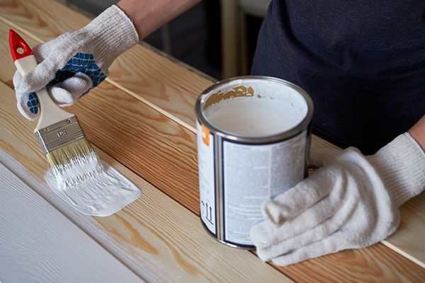 Person in gloves paints wood planks white with a brush and open paint can.