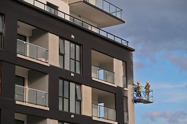 Workers in yellow hard hats on a lift painting a modern multi-story building exterior.