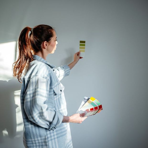 Woman holding paint swatches, comparing colors on a white wall.