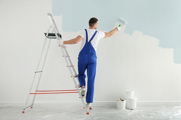 Person in blue overalls paints a wall blue, standing on a ladder; paint cans on the floor.