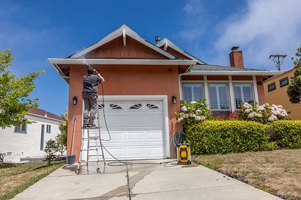 Person on ladder pressure washing a peach-colored house with a white garage door, under a blue sky.