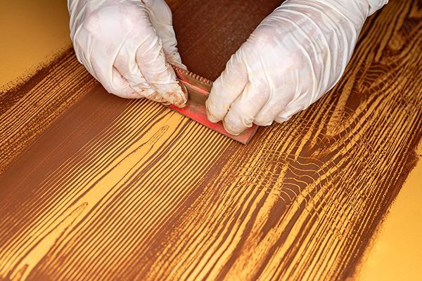 Hands in gloves using a tool to apply stain to wooden planks.