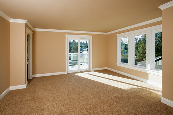 Empty room with tan walls, carpet, and white trim; sliding glass door and windows overlook trees.