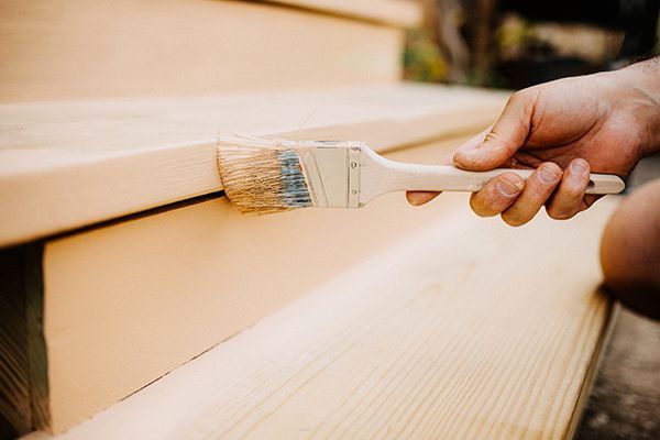 Person's hand using a paintbrush to apply stain to wooden outdoor steps.
