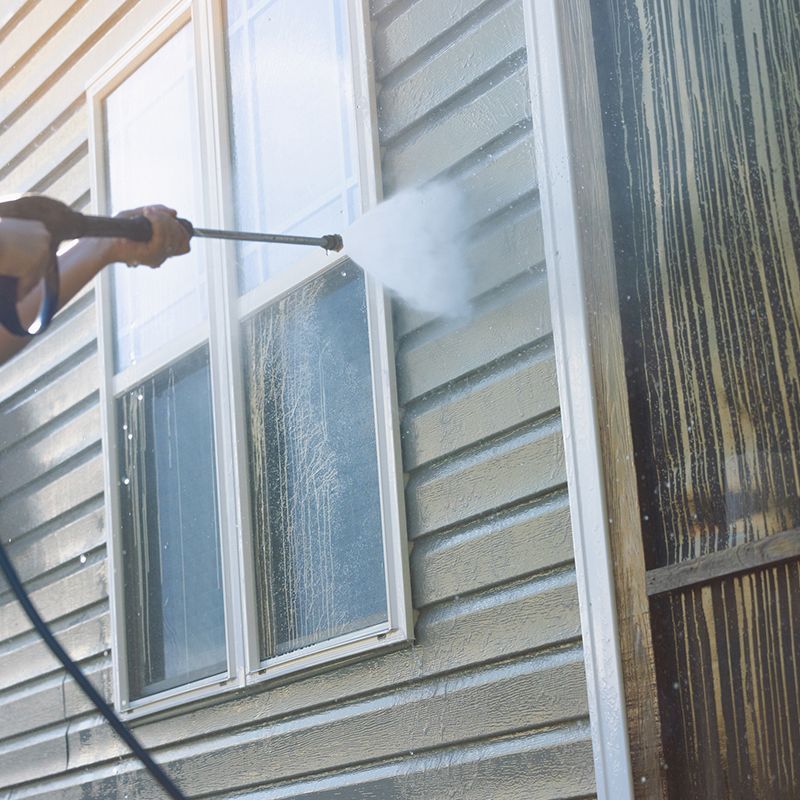 Person using a pressure washer to clean siding near a window. Dirty streaks on the siding are visible.