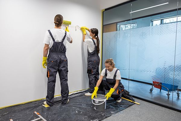 Three people painting a white wall in an office, wearing overalls and gloves.