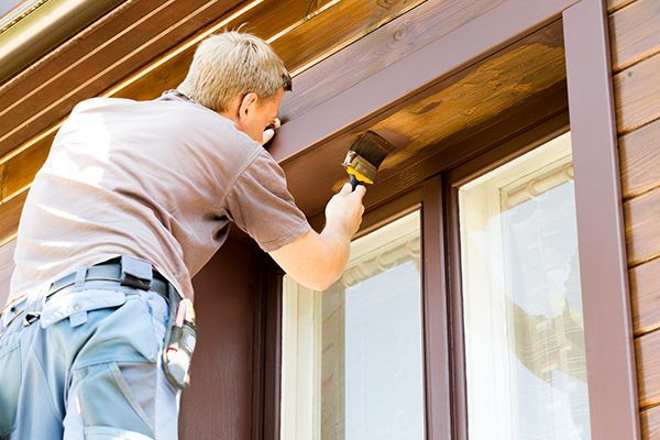 Man painting brown trim around a window on a wooden building exterior.