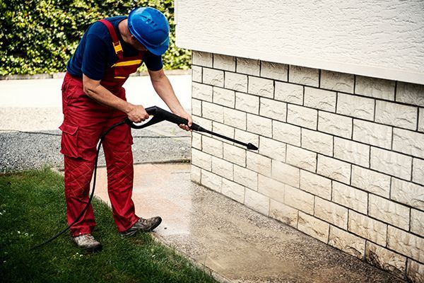 Man in red overalls and blue hard hat power washing a brick wall outside.