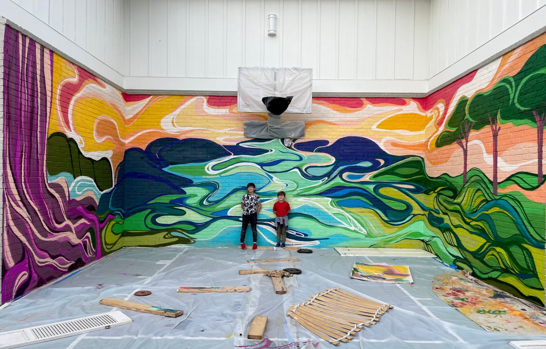 Two children stand in a room with colorful, mosaic-like landscape murals on the walls. A basketball hoop is visible.