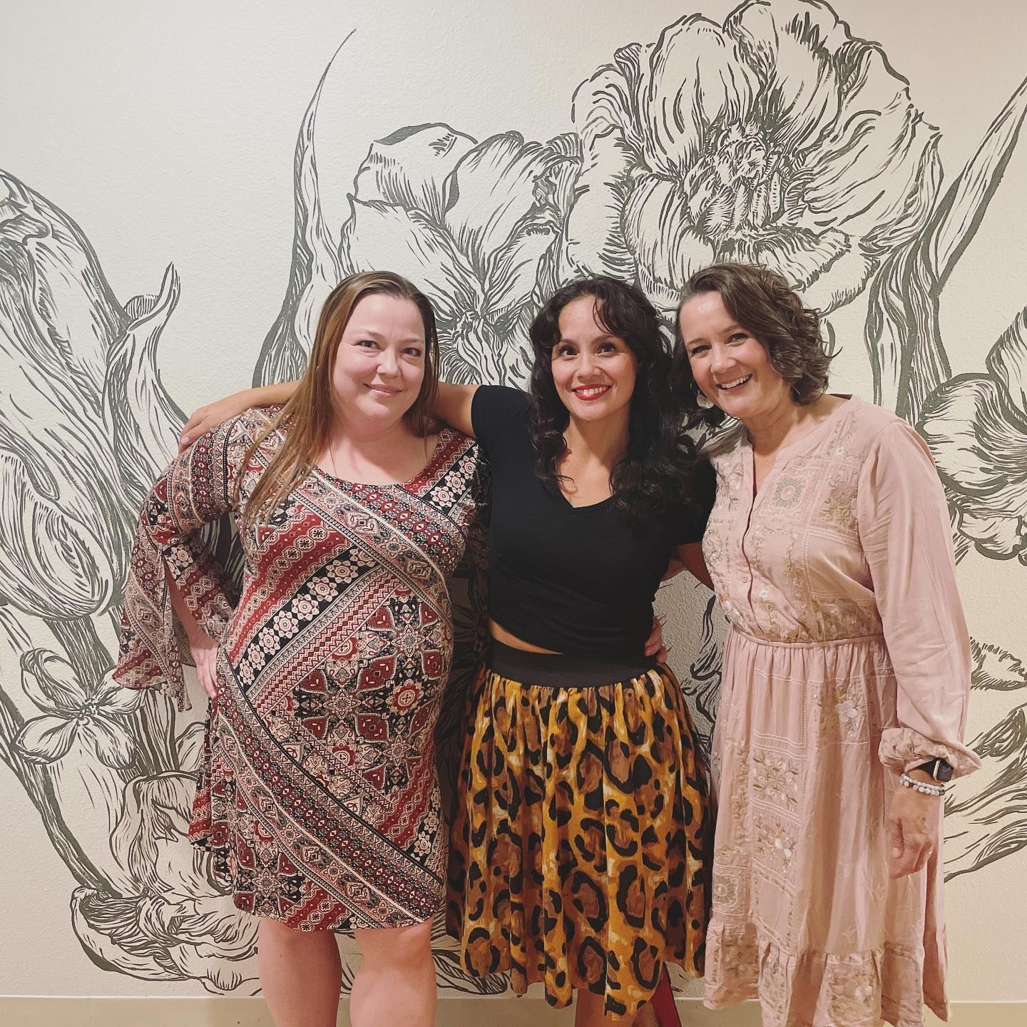 Three women smiling and posing in front of a floral wall mural.