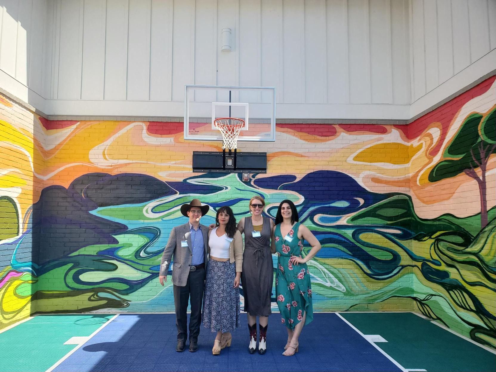 Four people pose on a basketball court with a colorful mural backdrop; they smile at the camera.