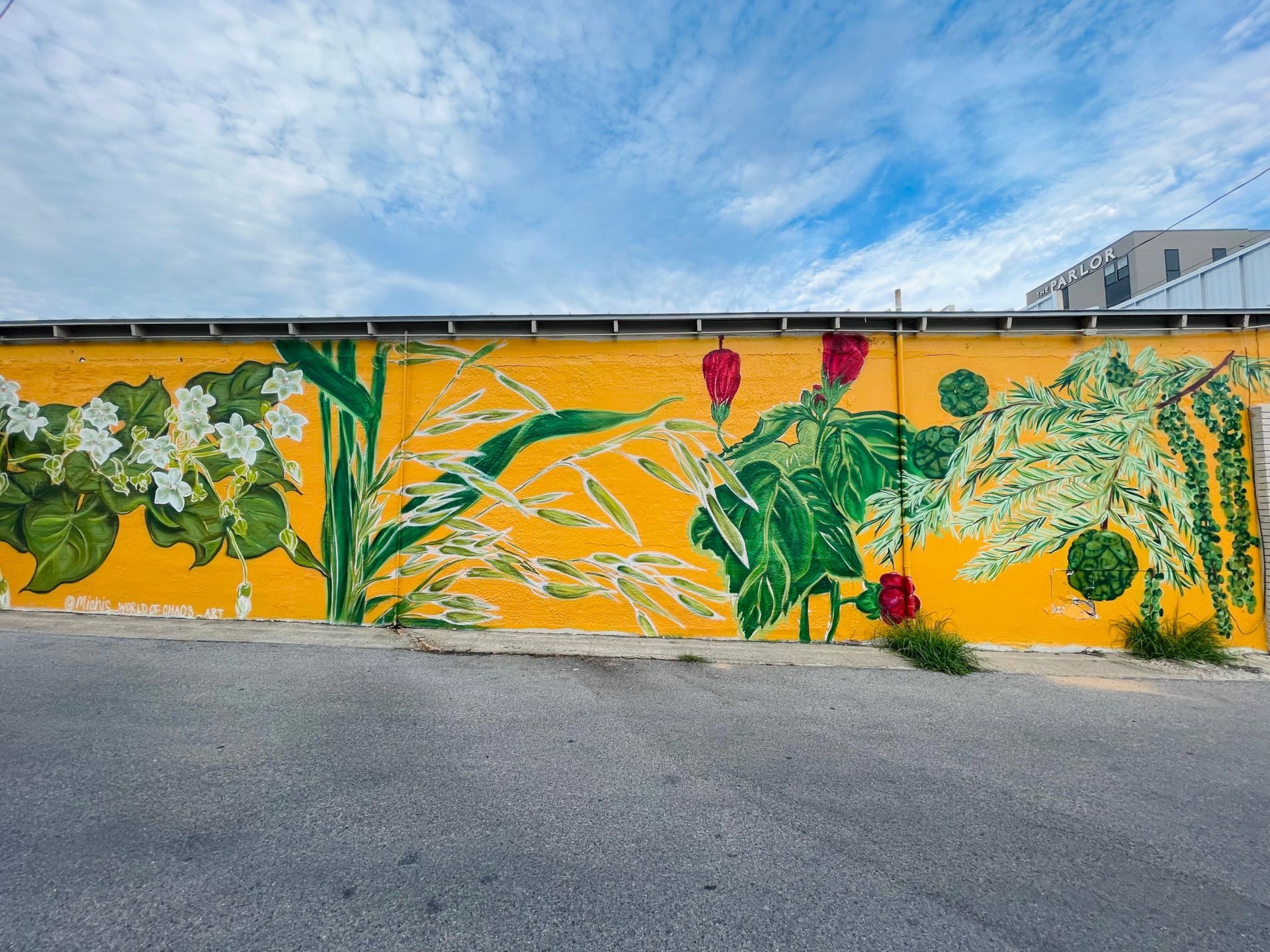Yellow mural with vibrant green and red plant illustrations on a building exterior under a partly cloudy sky.