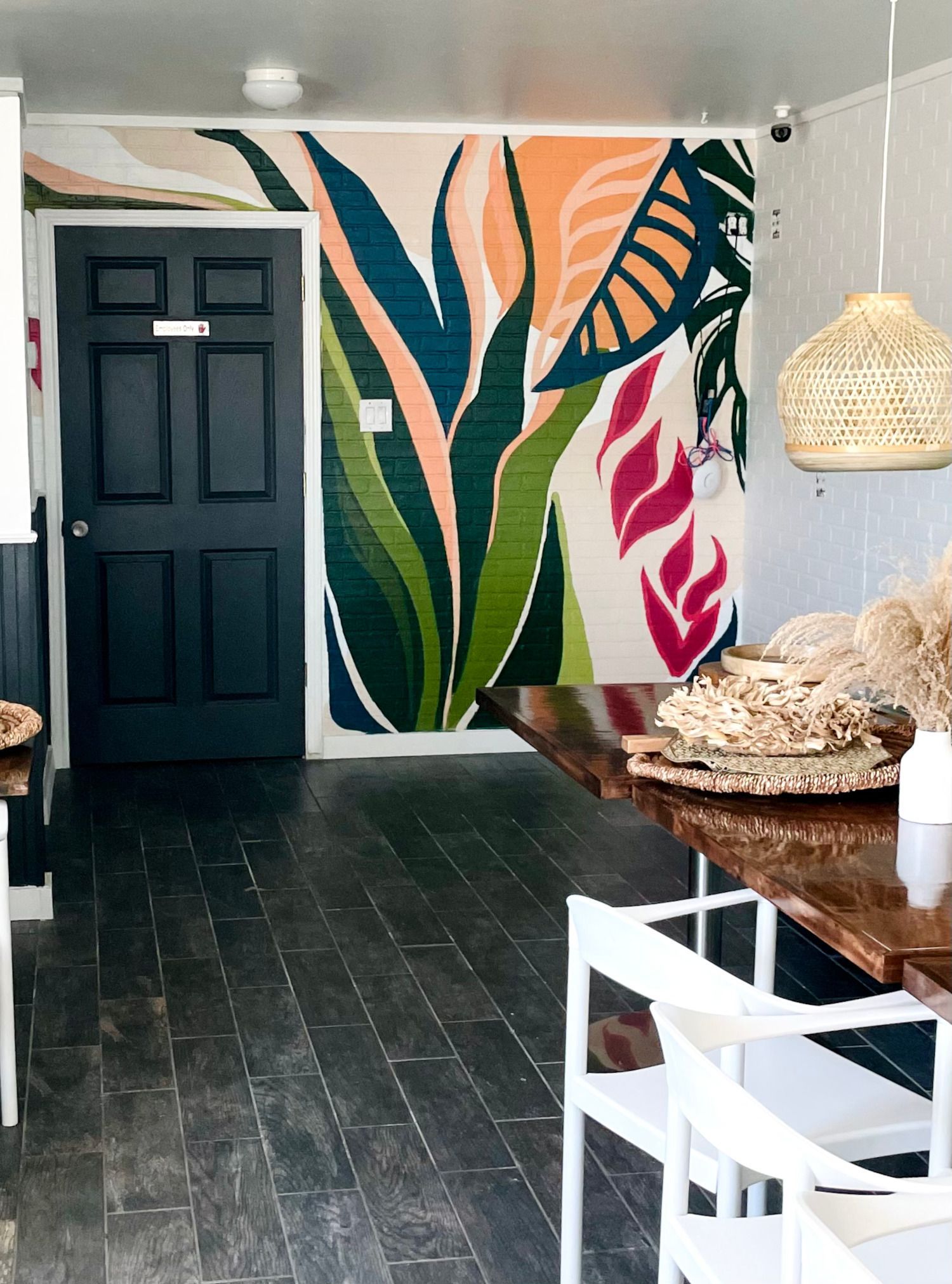 Entryway with a colorful botanical mural, dark door, wood table with white chairs, and woven light fixture.