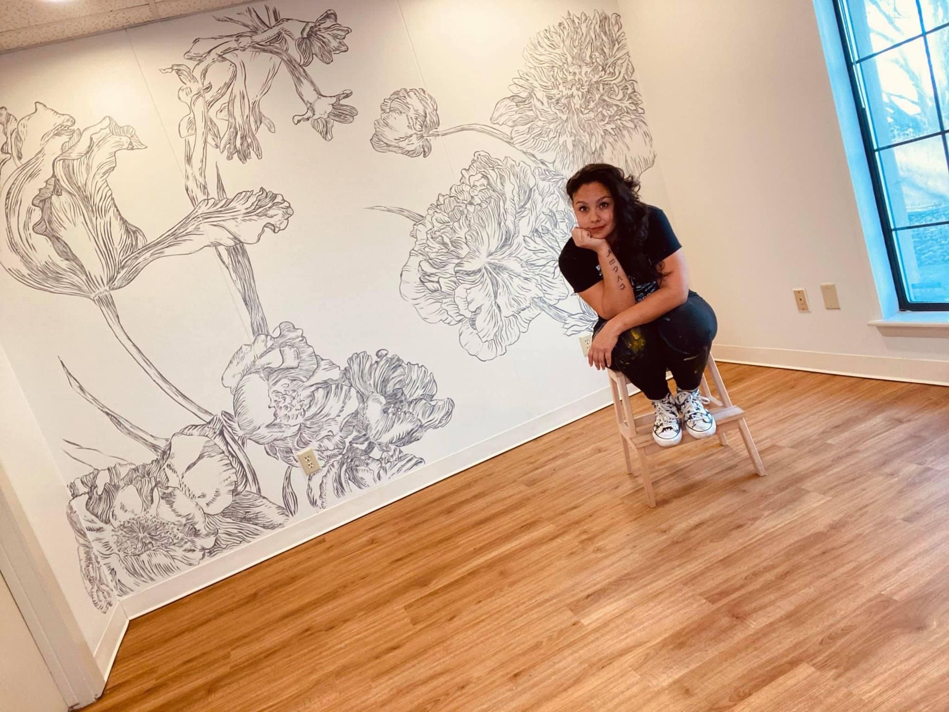 Woman poses next to a floral mural she created on a white wall; wooden floor, window.