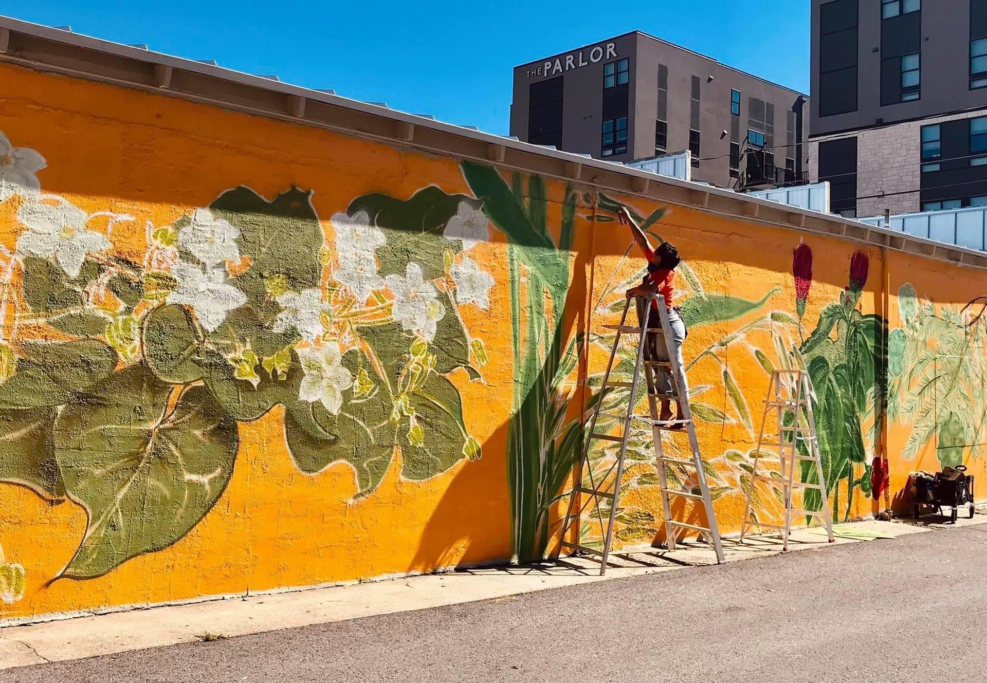 Artist on a ladder paints a floral mural on a yellow wall, with buildings in the background.