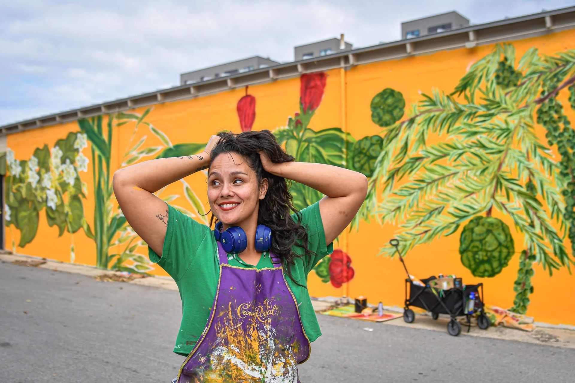Woman with headphones and paint-splattered apron poses in front of a colorful mural of plants.