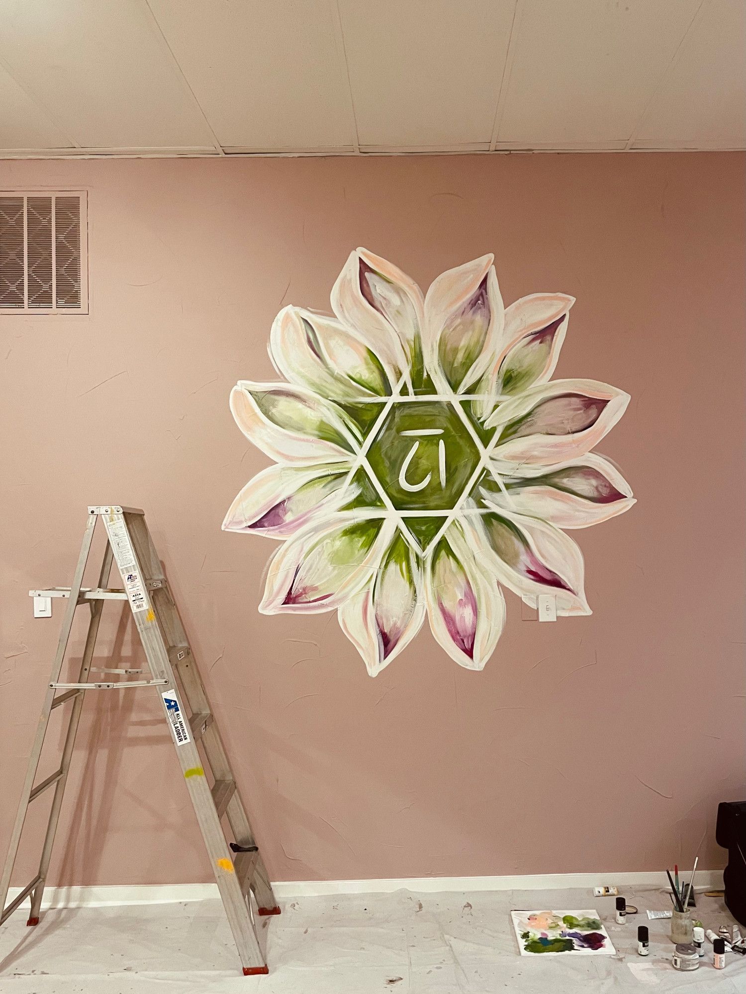 Wall mural of a flower mandala being painted, with a ladder and paint supplies in a pink-walled room.
