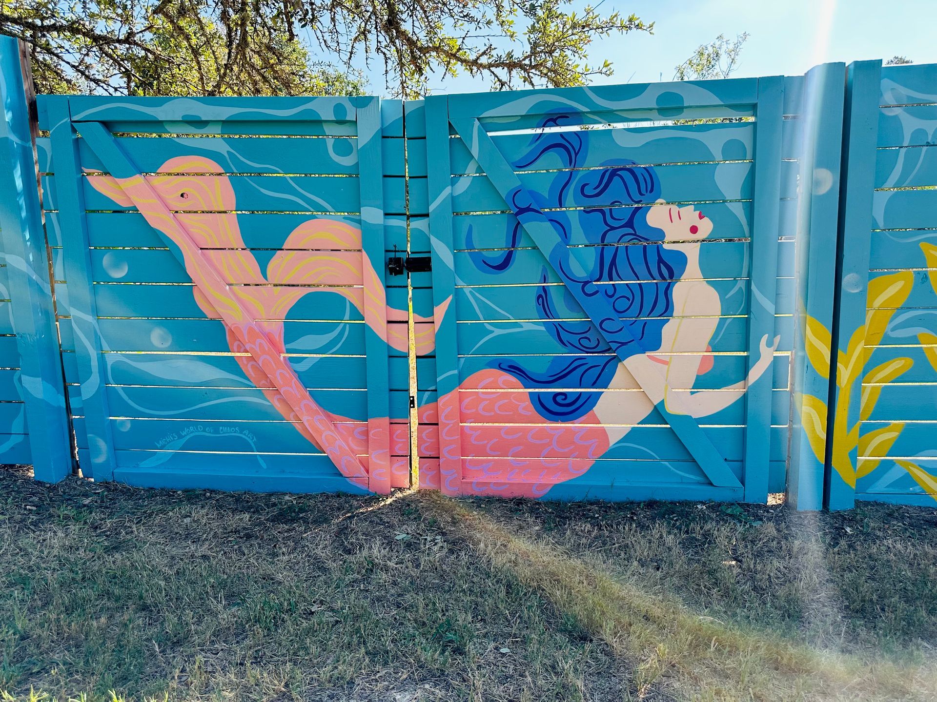 Mermaid painted on a turquoise fence with blue hair, pink tail, and yellow seaweed.
