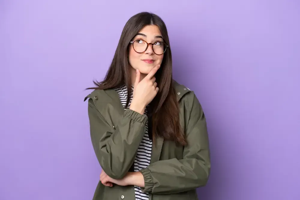 A young woman wearing glasses is thinking and looking up against a purple background.