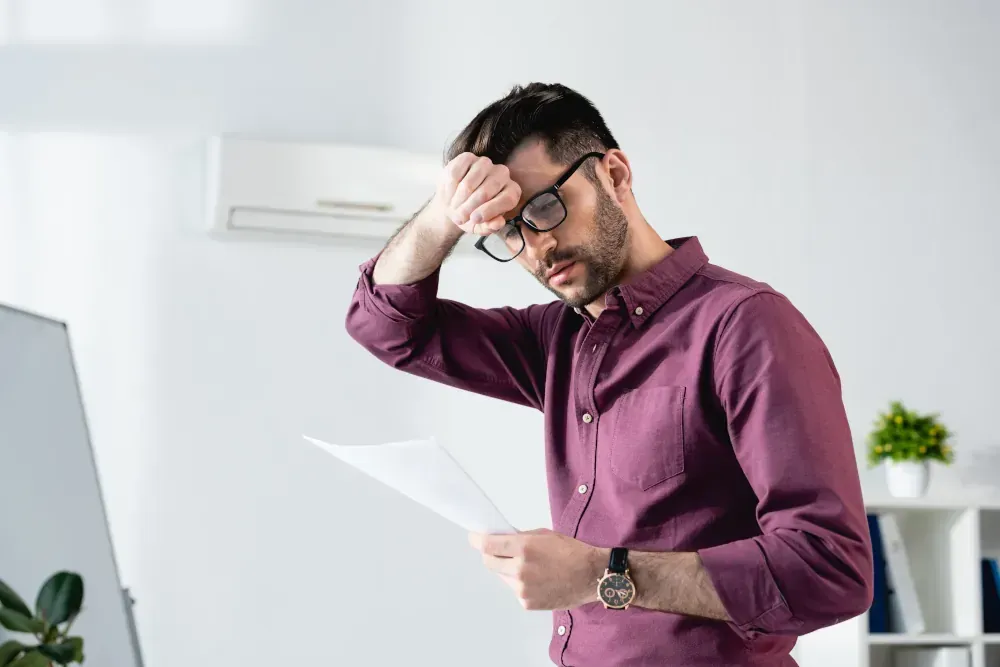 A man wearing glasses is holding his head while looking at a piece of paper.