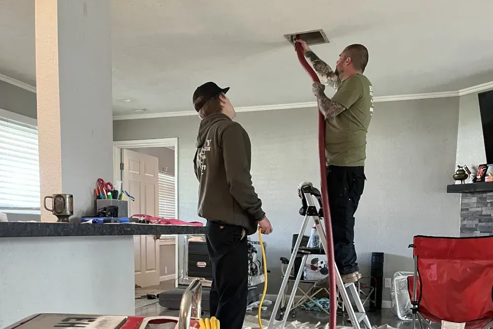 Two men are working on a ceiling vent in a living room.
