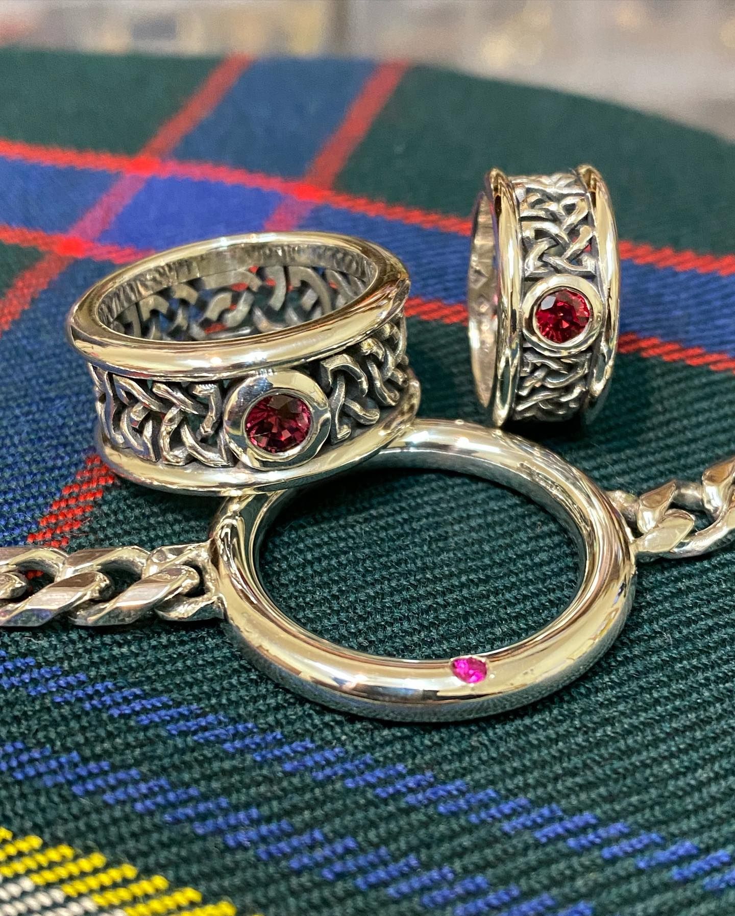 Three silver rings with small red gemstones and intricate knotwork designs resting on a blue, green, and red plaid fabric — Burchell's Jewellers In Bowral, NSW