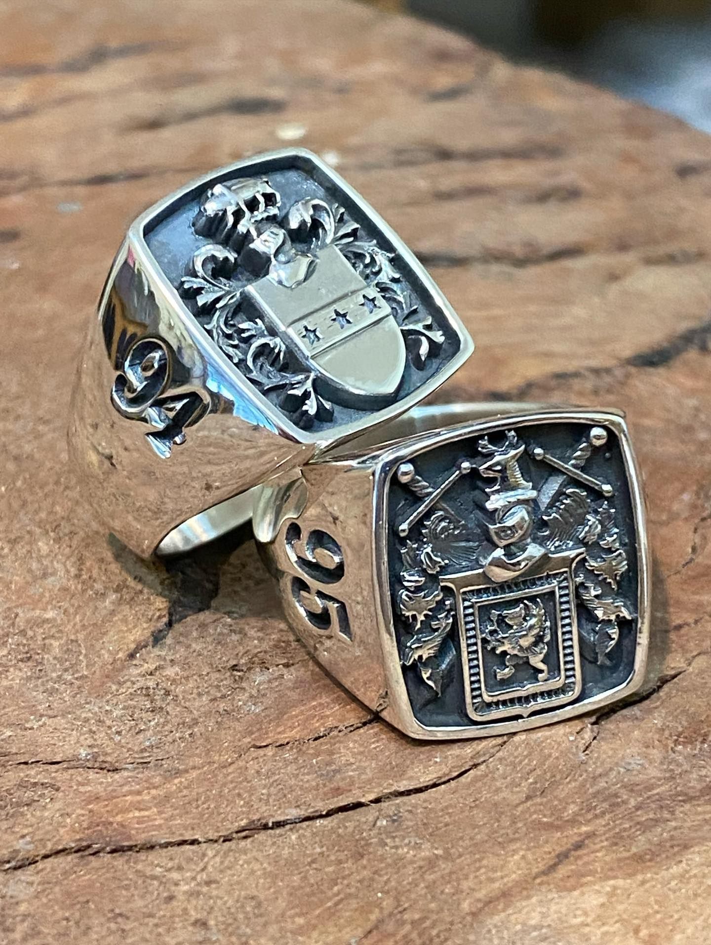 Two silver signet rings with engraved crests and the numbers 94 and 95, resting on a textured wooden surface — Burchell's Jewellers In Bowral, NSW