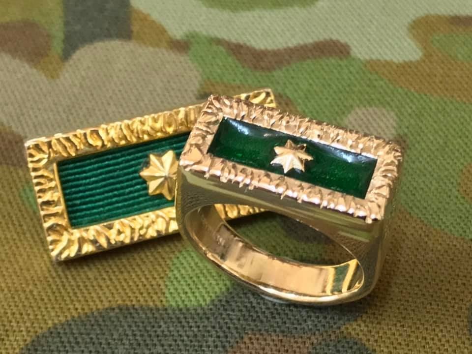 A gold ring and a matching pin, both featuring a dark green rectangular center with a small gold star, on camo fabric. — Burchell's Jewellers In Bowral, NSW