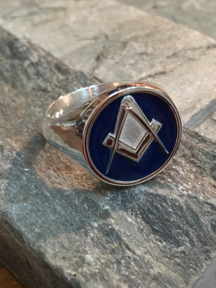 A silver ring with a blue circular face featuring the gold Masonic Square and Compasses symbol, resting on a stone ledge — Burchell's Jewellers In Bowral, NSW