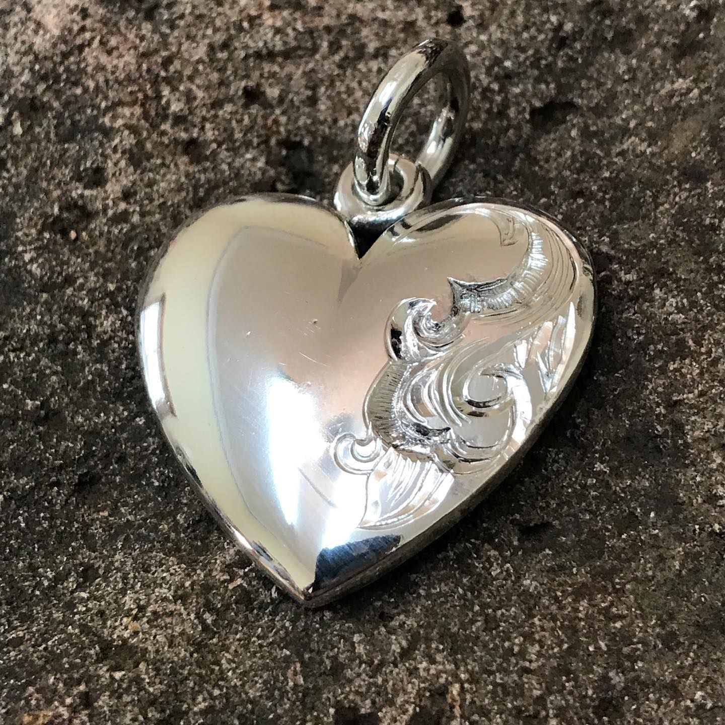 A shiny silver heart pendant with an etched scrollwork design on the right side, resting on a rough, dark surface — Burchell's Jewellers In Bowral, NSW