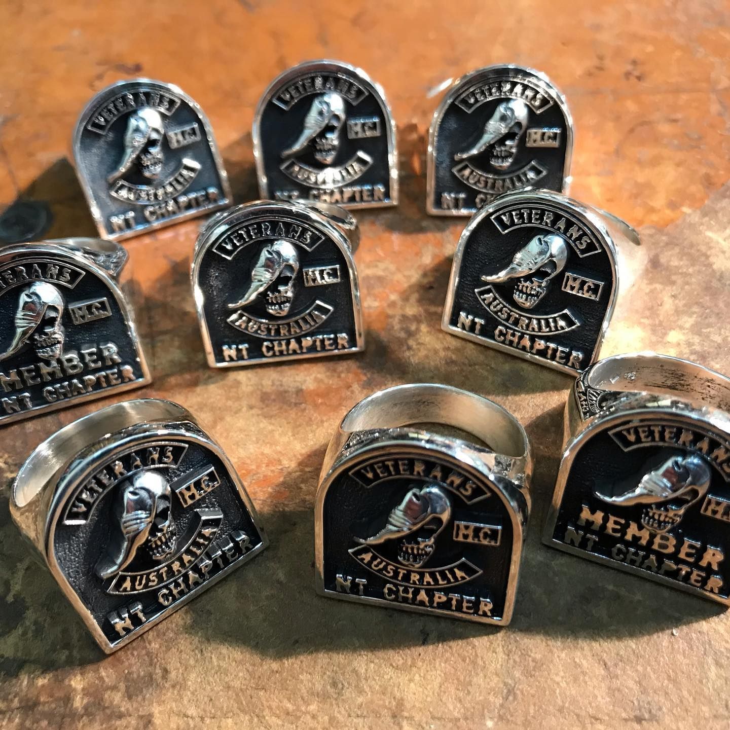 A collection of nine silver Veterans MC Australia, NT Chapter rings arranged on a textured wooden surface. — Burchell's Jewellers In Bowral, NSW
