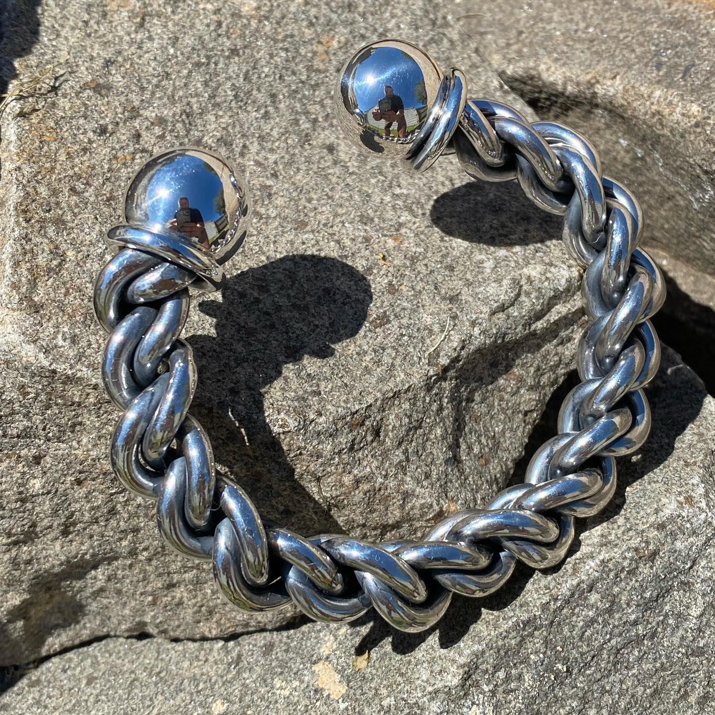 A silver-toned, braided metal cuff bracelet with spherical ends, resting on a textured, grey stone surface — Burchell's Jewellers In Bowral, NSW