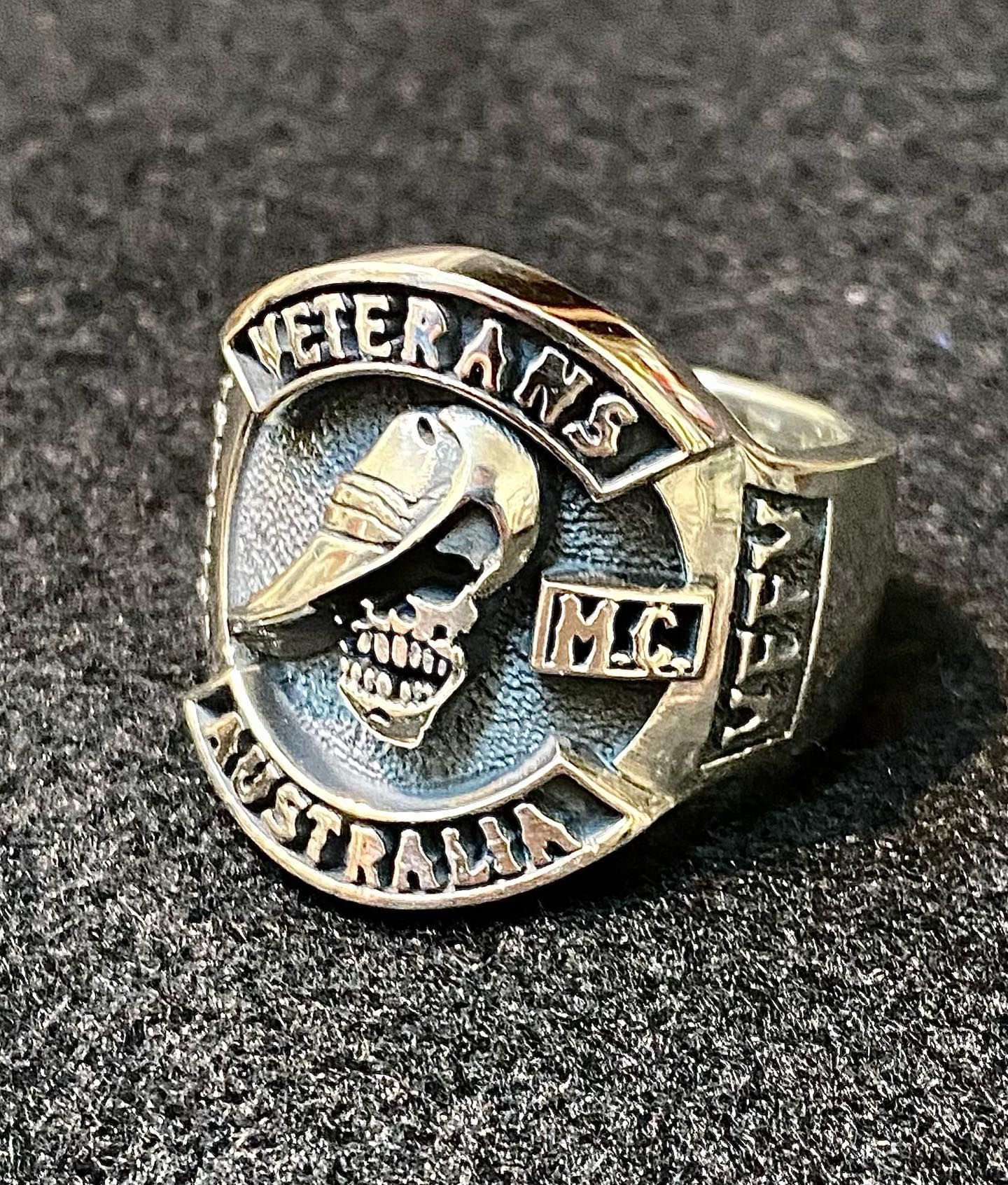 A metallic Veterans MC Australia ring featuring a skull wearing a hat, set against a dark, textured background — Burchell's Jewellers In Bowral, NSW