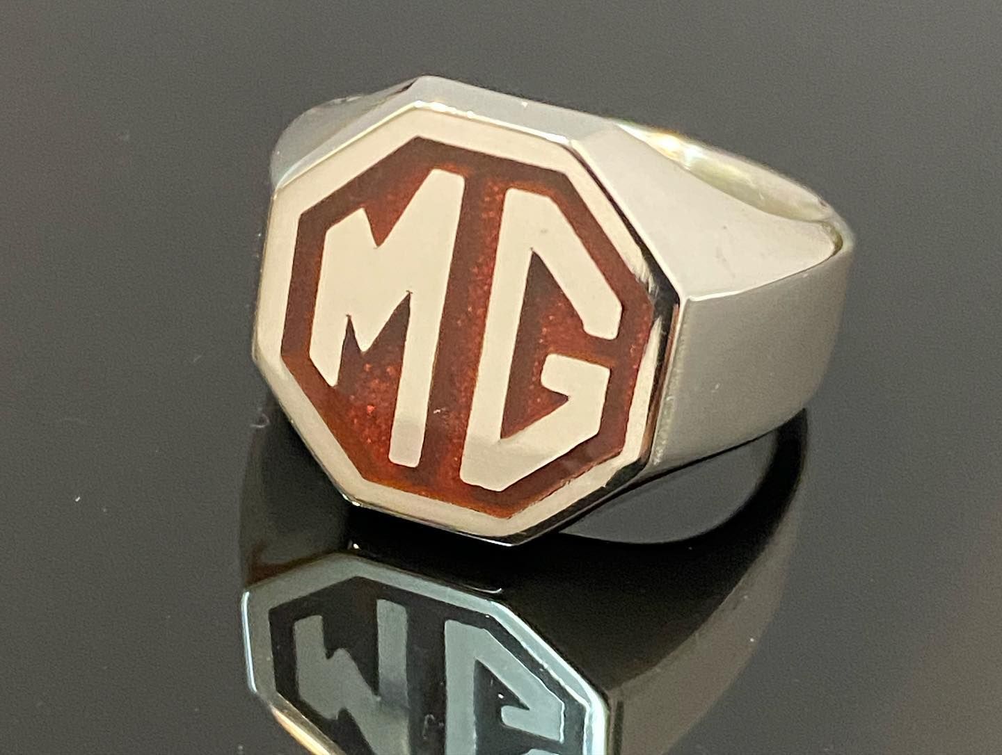 A silver ring with an octagonal face featuring the maroon and silver MG logo, reflecting on a dark, glossy surface — Burchell's Jewellers In Bowral, NSW