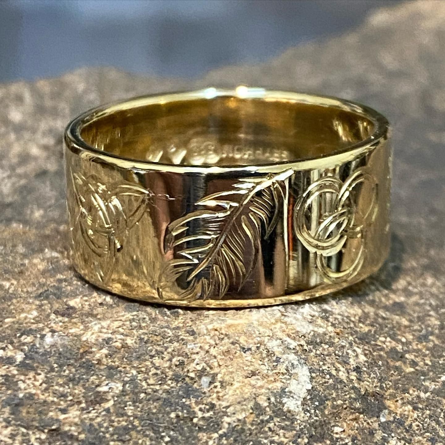 A gold ring with engraved feather and Celtic knot designs, resting on a textured, stony surface. — Burchell's Jewellers In Bowral, NSW