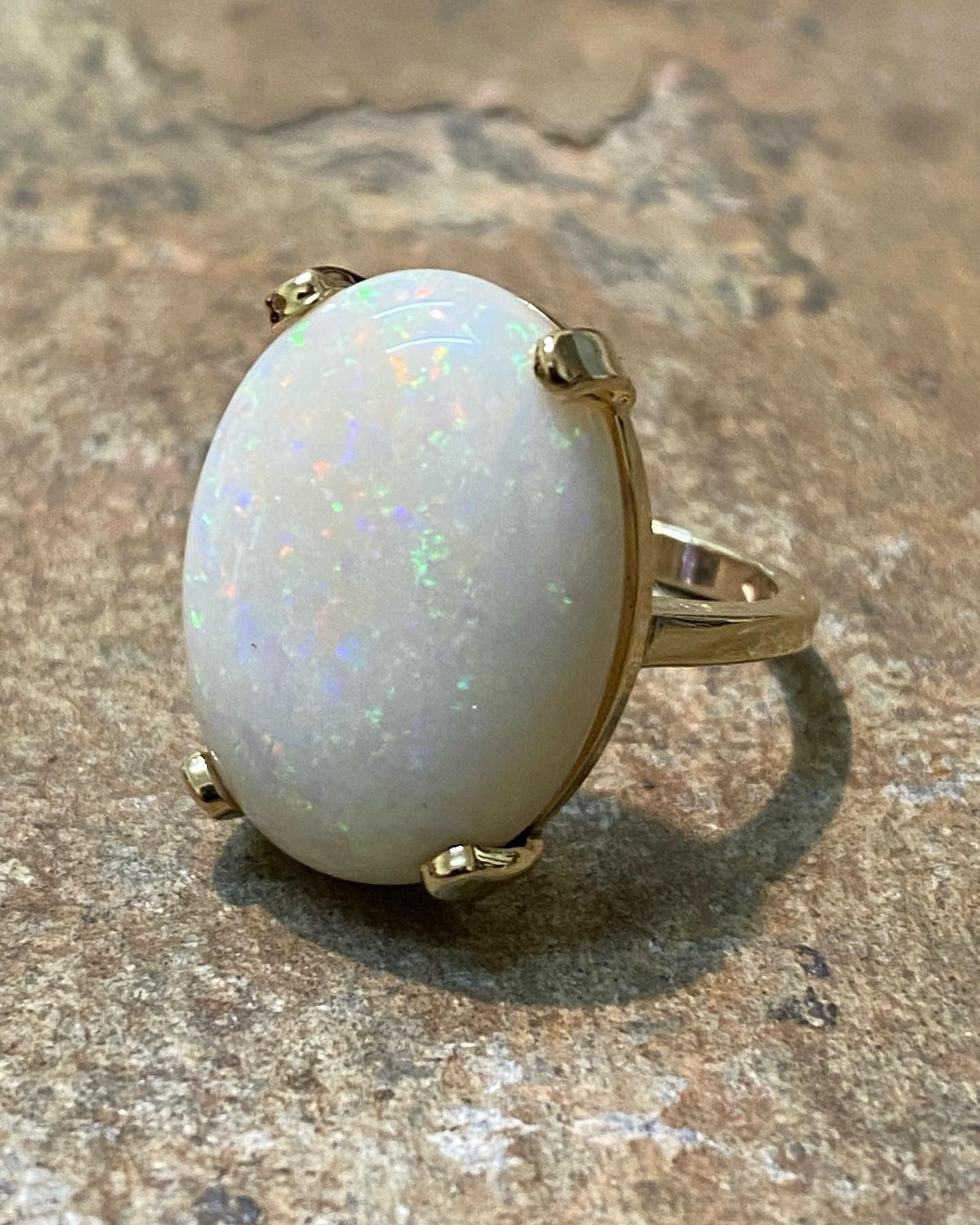 An oval-shaped, iridescent white opal gemstone set in a gold ring, resting on a textured, light brown stone surface. — Burchell's Jewellers In Canberra, NSW
