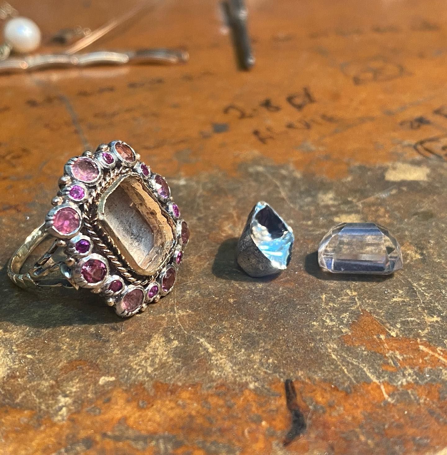 An empty decorative ring with pink stones rests on a wooden surface next to a loose grey stone and a faceted crystal. — Burchell's Jewellers In Bowral, NSW