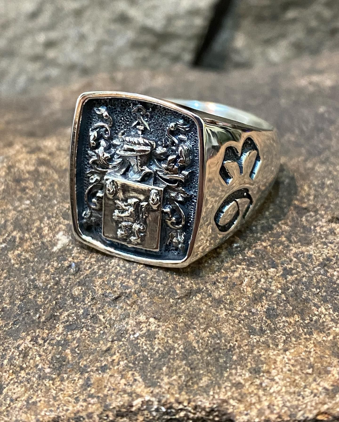 A silver signet ring with an engraved family crest and ornate side designs, resting on a textured, rough stone surface — Burchell's Jewellers In Bowral, NSW
