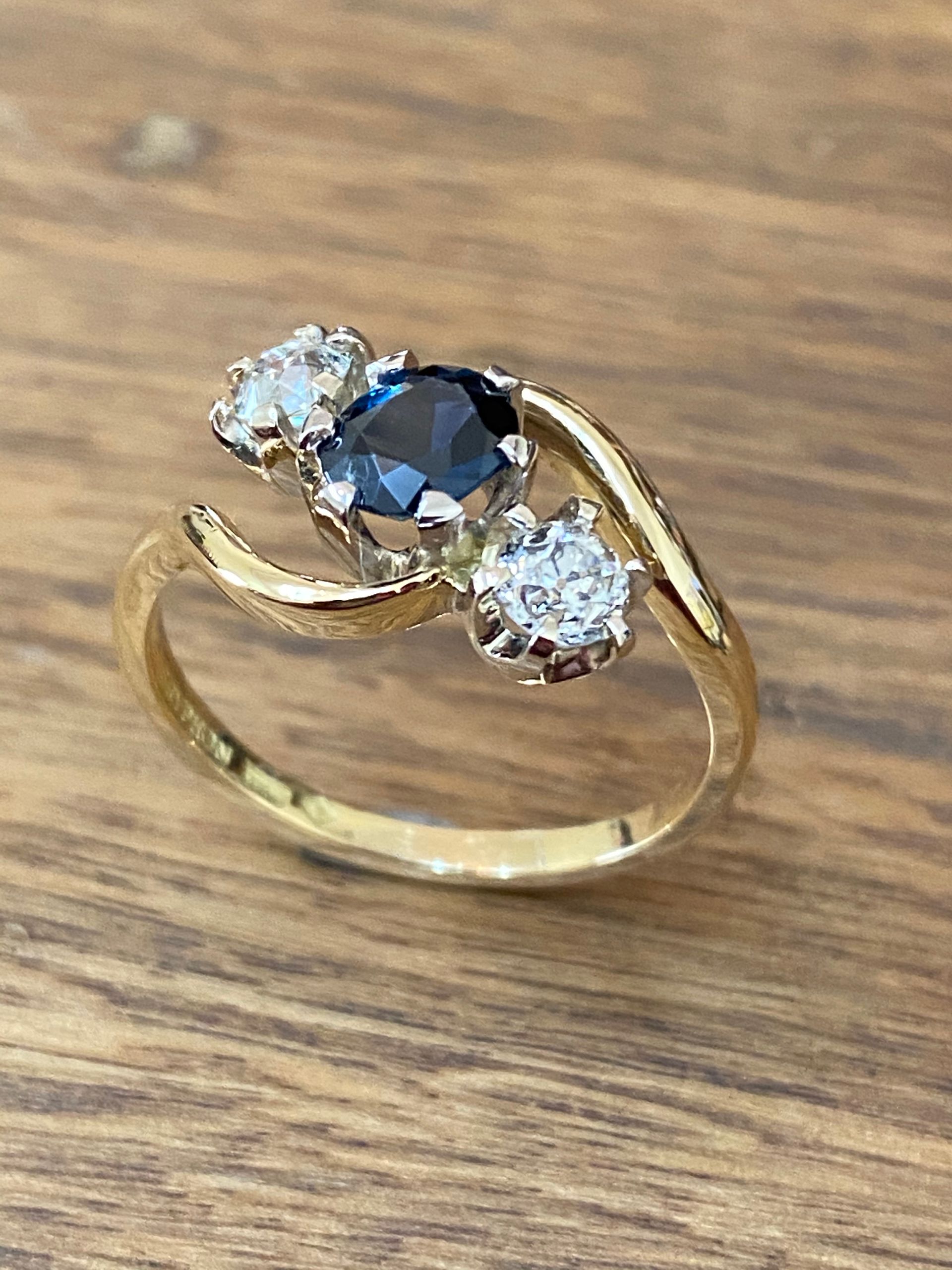 Gold Ring With a Blue Gemstone and Two White Gemstones Set on a Wooden Surface — Burchell's Jewellers In Bowral, NSW