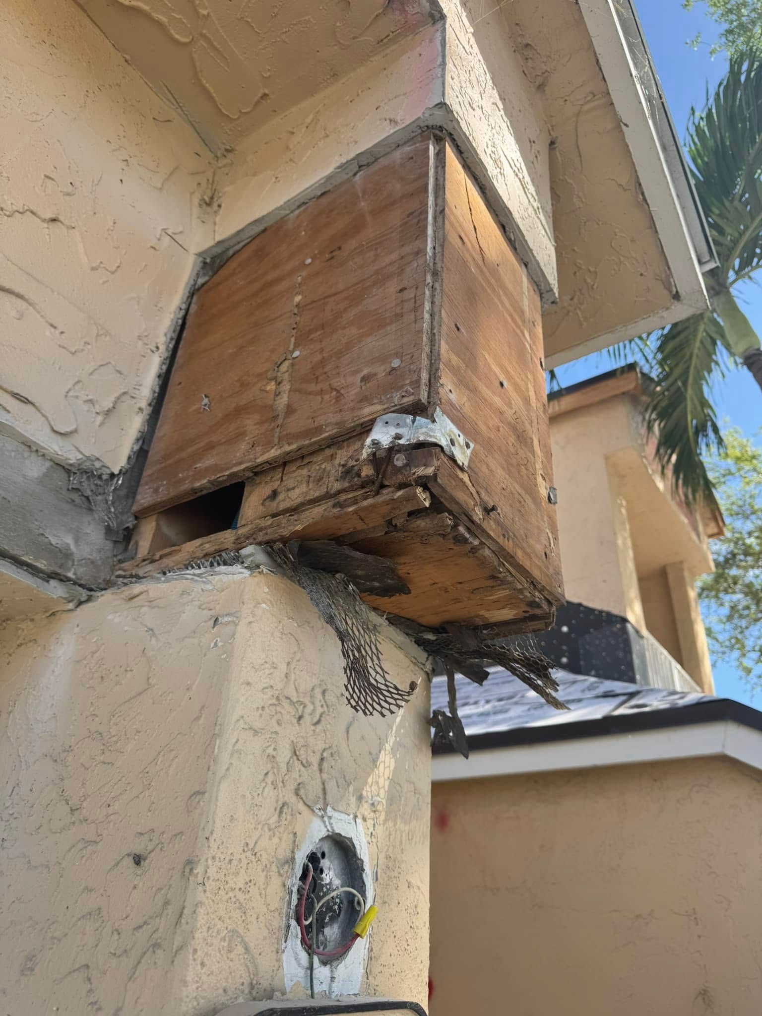 Damaged exterior wall corner with exposed wooden framing and debris near an outdoor electrical fixture.