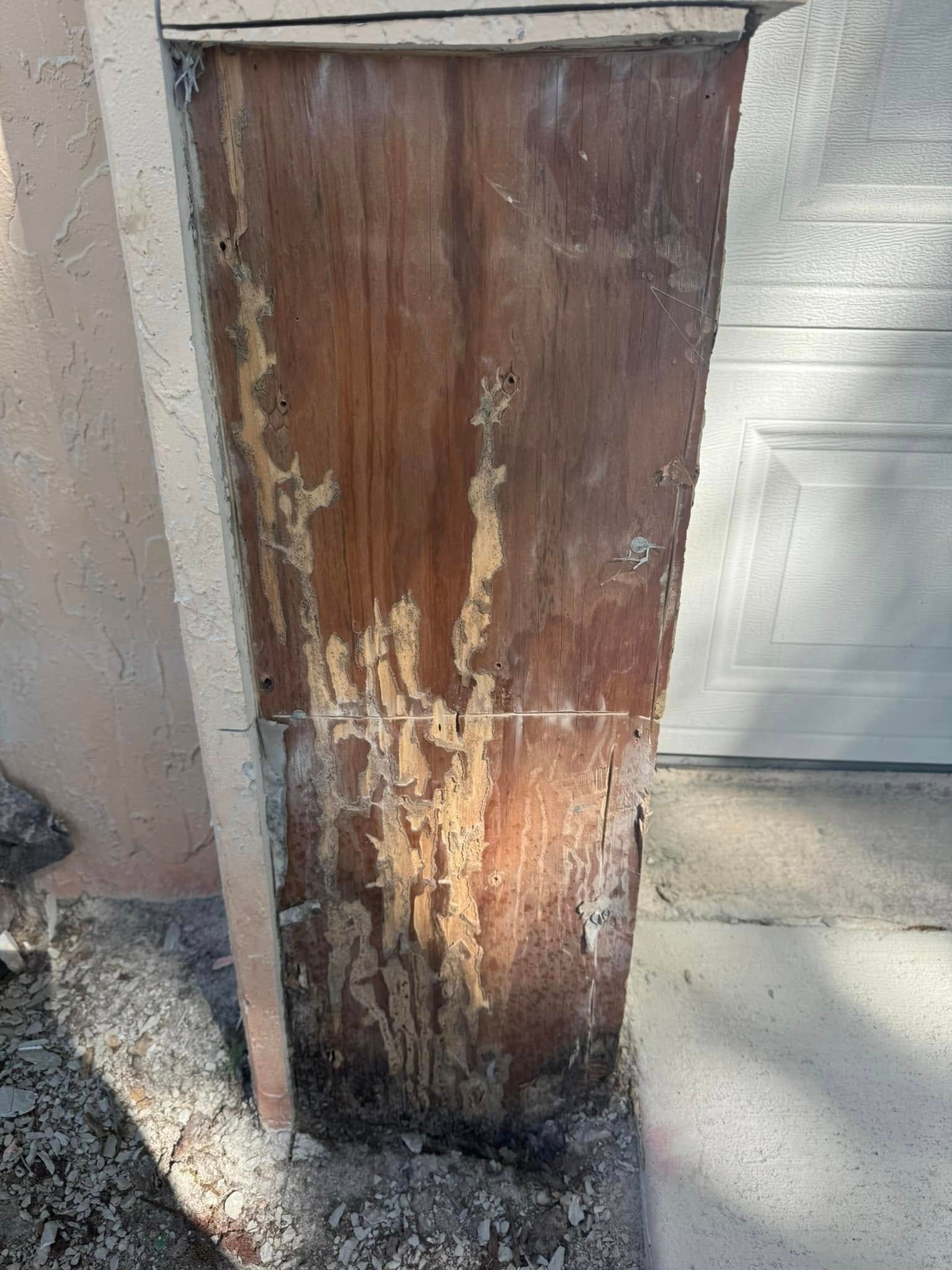 A severely weathered wooden pillar with visible surface damage and decay, situated next to a house wall and garage door.