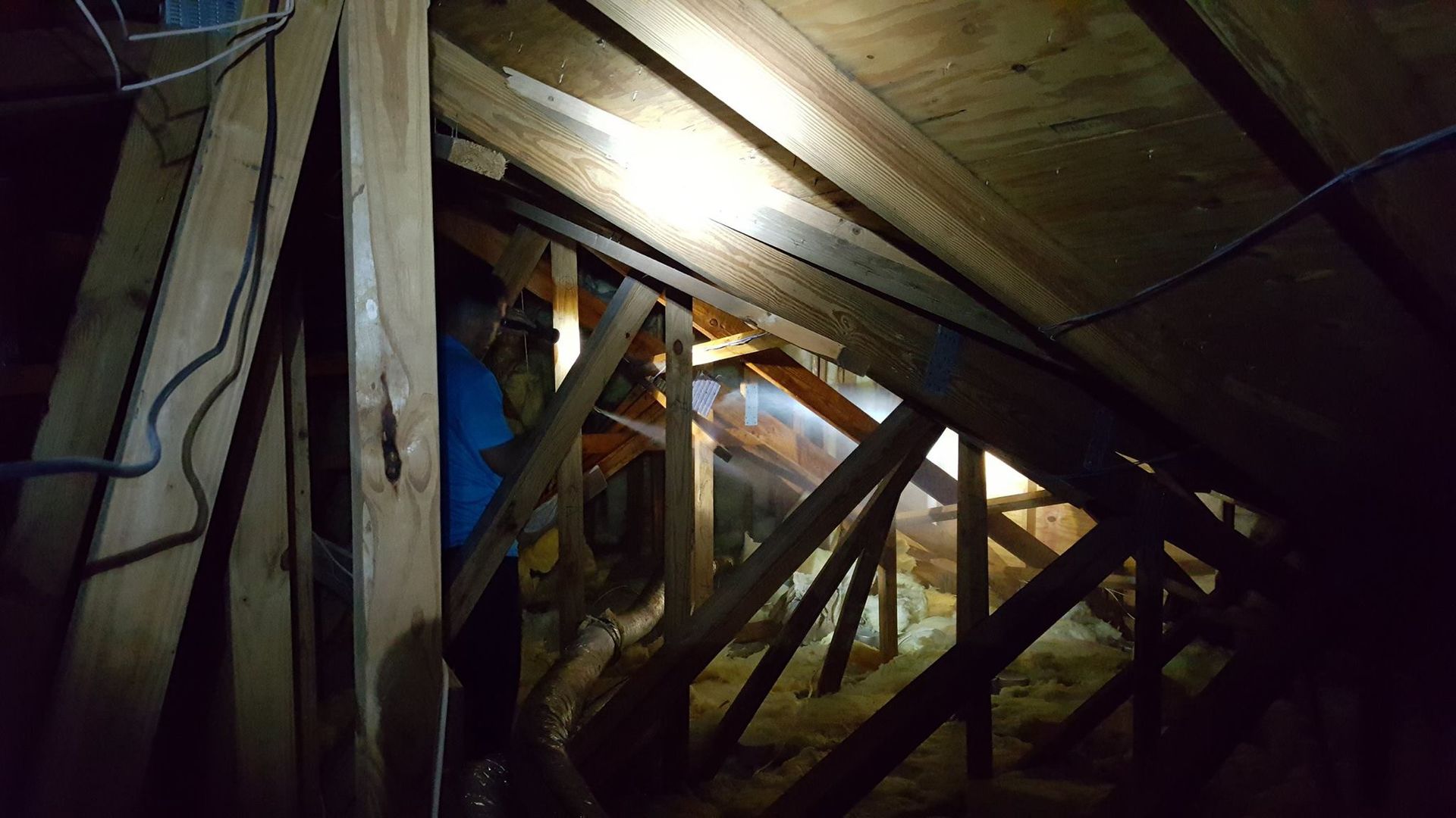 A person in an attic inspecting wooden roof rafters, seen in the dim light of a flashlight.