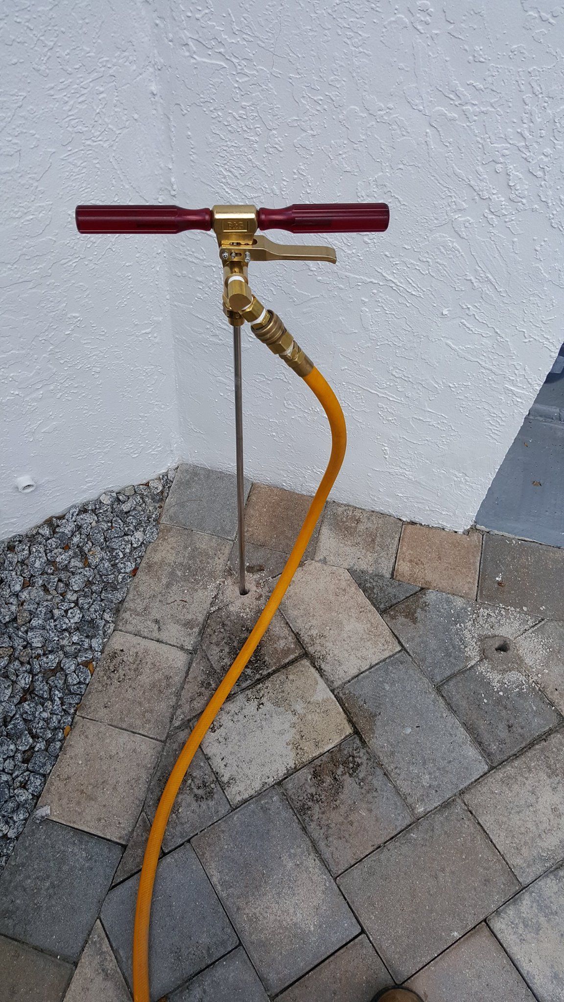 A brass probe tool with a red handle and an attached yellow hose is inserted into a crack in outdoor paving stones.
